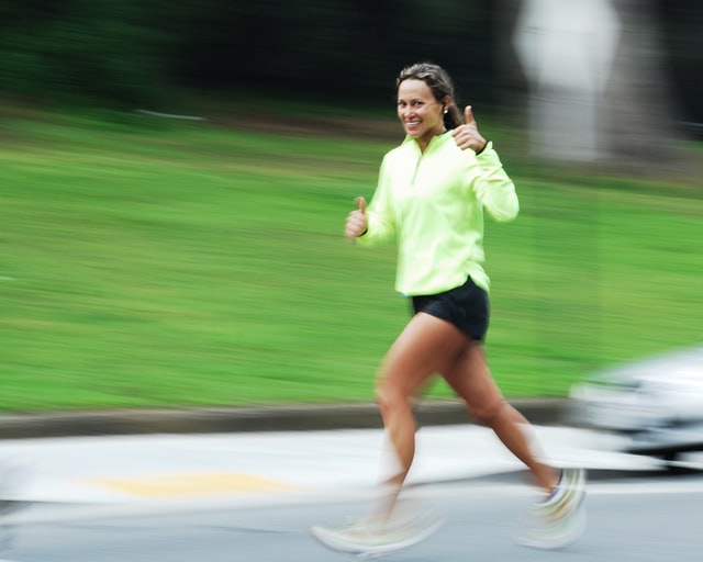 Foto einer sportlichen wirkenden jungen Frau, die im Vorbei-Joggen lächelnd einen Daumen nach oben hält.
